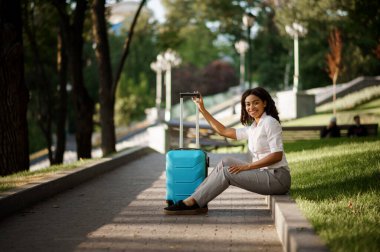 Yaz parkında elinde bavuluyla oturan genç bir kadın. Bagajı açık havada gezen bayan yolcu, çantalı yolcu doğada dinleniyor. Bagajı olan kız şehir sokağında rahatlar.