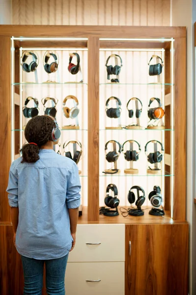 音響店でヘッドフォンを選択する女性 バックビュー オーディオショップで女性の人 背景にイヤホンでショーケース マルチメディアサロンで購入者 — ストック写真