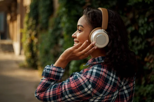 夏の公園で音楽を聴いてヘッドフォンで陽気な女性 女性音楽ファン屋外を歩く イヤホンの女の子 背景に緑の茂み — ストック写真