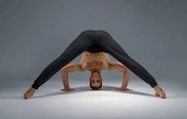 Male yoga stands on his head, hands and legs, meditation, grey background. Strong man doing yogi exercise, asana training, top concentration, healthy lifestyle
