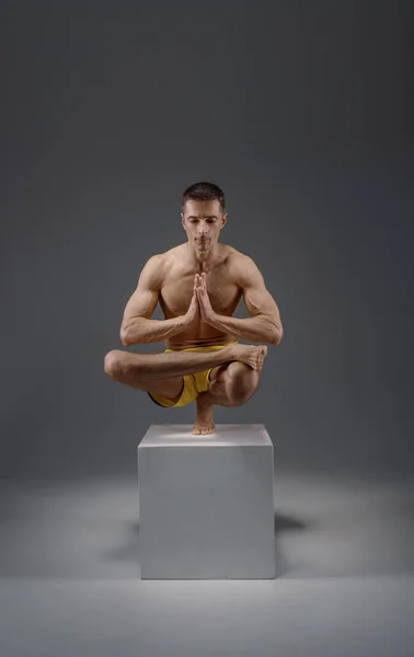 Male yoga keeps balanc on hands in difficult pose on the pedestal, meditation position, grey background. Strong man doing yogi exercise, asana training, top concentration, healthy lifestyle