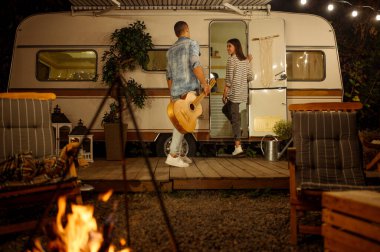 Gitarlı genç bir çift ormanda kamp yaparken romantik piknik yapıyor. Karavanda yaz macerası, arka planda kamp arabası. Erkek ve kadın şenlik ateşinin yanında, karavanla seyahat ediyor.