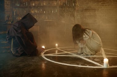 Siyah kapüşonlu erkek şeytan çıkarıcı bir kadından şeytanı kovuyor. Şeytan çıkarma, gizemli paranormal ayin, karanlık din, gece korkusu, arka planda raflarda duran iksirler.