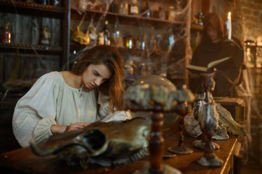 Siyah kapüşonlu erkek şeytan çıkarıcı korkunç kadından iblisleri kovuyor. Şeytan çıkarma, gizemli paranormal ayin, karanlık din, gece korkusu, arka planda raflarda duran iksirler.