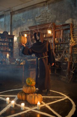 Siyah kapüşonlu erkek şeytan çıkarıcı balkabağını sihirli çemberin yakınında tutuyor. Şeytan çıkarma, gizemli paranormal ayin, karanlık din, gece korkusu, arka planda raflarda duran iksirler.