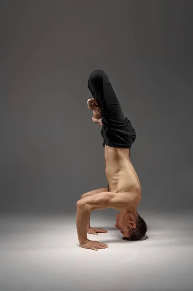 Male yoga standing on his head and hands, meditation, grey background. Strong man doing yogi exercise, asana training, top concentration