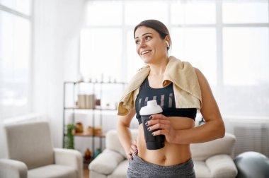 Eğitimden sonra kadın elinde su şişesi tutuyor. Spor giyim, internet sporları, arka planda oda içi.