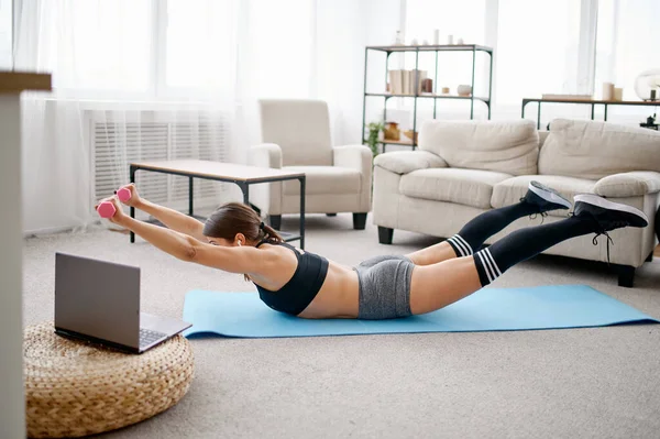 Woman doing abs exercise with dumbbells, online fitness training at the laptop. Female person in sportswear, internet sport workout, room interior on background