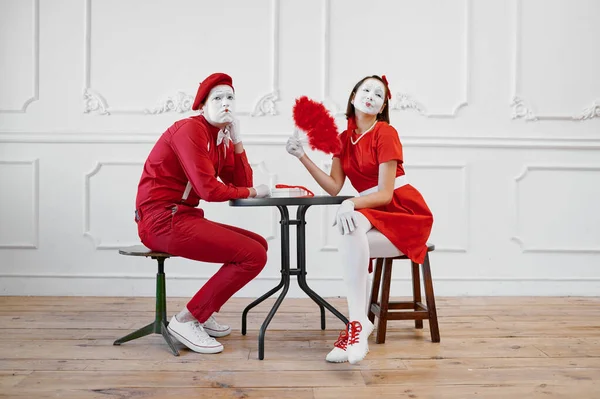 Two mime artists in red costumes, scene at the table. Pantomime theater, parody comedian, positive emotion, humour performance, funny face mimic and grimace