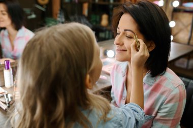 Sevimli kız, makyaj salonunda annesine makyaj yapıyor. Anne ve küçük kız birlikte oyun stilistleri, mutlu çocukluk, büyüleyici aile.