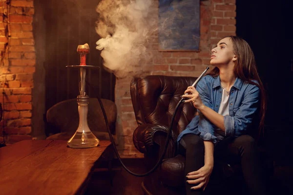 Young woman blows smoke in hookah bar, chill out. Shisha smoking, traditional bong culture, tobacco aroma for relaxation, rest with hooka