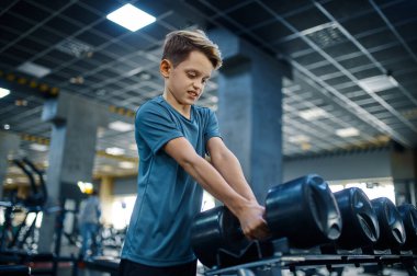 Çocuk spor salonunda ağır dambıl almaya çalışıyor. Spor kulübünde eğitim gören genç, sağlık hizmetleri ve sağlıklı yaşam tarzı, spor yapan okul çocuğu.