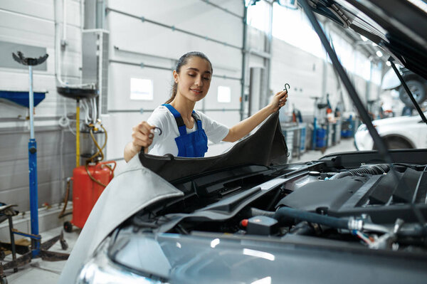 Female mechanic stands at the hood, car service. Vehicle repairing garage, woman in uniform, automobile station interior on background