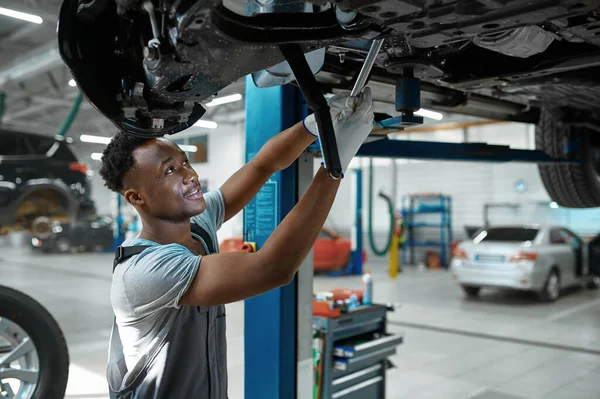 Male mechanic checks car suspension, auto service. Vehicle repairing garage, man in uniform, automobile station interior on background