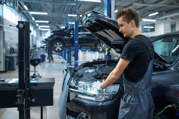 Male mechanic adjusts the headlights, car service. Vehicle repairing garage, man in uniform, automobile station interior on background