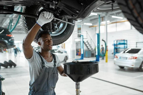 Male mechanic drains the oil, car service. Vehicle repairing garage, man in uniform, automobile station interior on background