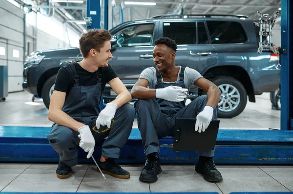 Two male mechanics sitting on car lift in auto service. Vehicle repairing garage, men in uniform, automobile station interior on background. Professional auto diagnostic