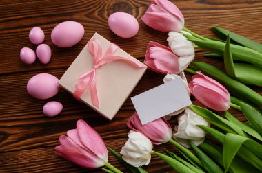 Pembe laleler, paskalya yumurtaları ve tahta arka planda hediye kutusu. Bahar çiçekleri ve paskalya yemekleri, taze çiçek süslemeleri, bayram kutlamaları sembolleri.
