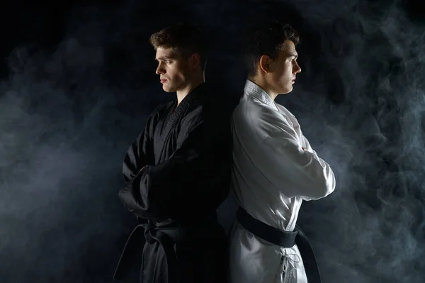 Two male karatekas in white and black kimono, dark background with smoke. Fighters on workout, martial arts, fighting competition