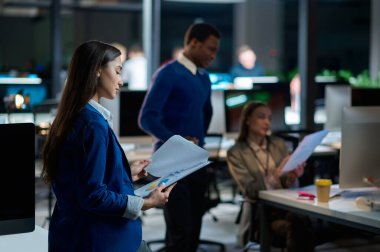 Genç yöneticiler gece ofisinde çalışıyor. Gülümseyen erkek ve kadın işçiler, arka planda karanlık iş merkezi, modern işyeri