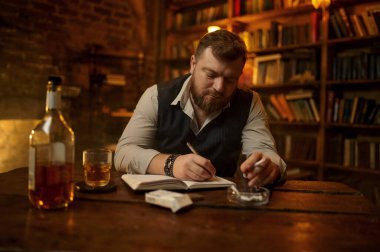 Adam sigara içiyor ve deftere yazıyor, arka planda ahşap masa var. Tütün içme kültürü. Erkek tiryaki.