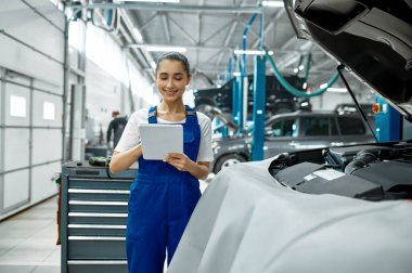 Dizüstü bilgisayarı olan kadın tamirci motor, araba servisi, profesyonel diyagtostik. Araç tamirhanesi, üniformalı kadın, arka planda otomobil istasyonu.