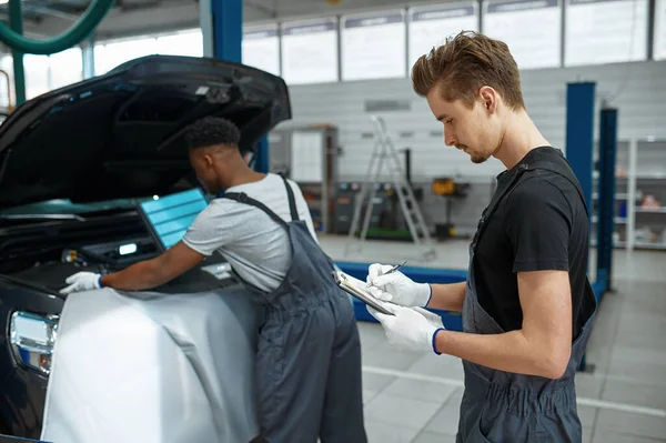 Two male workers repairs engine, car service. Vehicle repairing garage, men in uniform, automobile station interior on background. Professional auto diagnostic