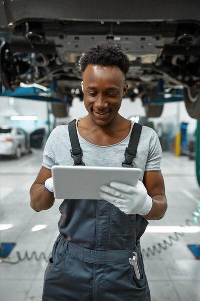 Работник с ноутбуком, машина на лифте, автосервис. Гараж для ремонта автомобилей, человек в форме, интерьер автовокзала на заднем плане
