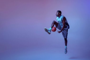 Basketbolcu stüdyoda topla antrenman yapıyor, neon arka planda. Spor giyim sektöründe profesyonel erkek oyuncu, sporcu.