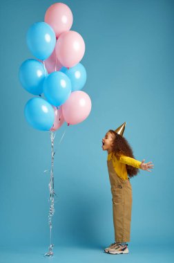 Şapkalı küçük kız elinde bir sürü balon tutuyor.