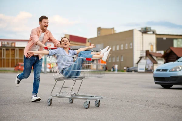 Funny family couple rides in cart on parking — Zdjęcie stockowe