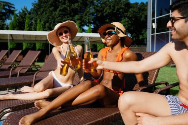 Arkadaşlar havuz kenarında bira şişelerini tokuştururlar.