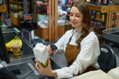 Bir kadın, elinde bir paket ve tarayıcıyla kasada süt tarıyor.