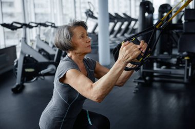 Yaşlı bir kadın spor salonunda spor yaparken süspansiyon antrenörü üzerinde çeşitli egzersizler yapmakla meşgul.