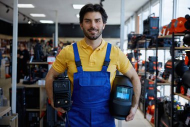 Mavi tulumlu işçi bir mağazanın içinde kaynak makinesi tutuyor. İşine bakıyor ya da hazırlanıyor.