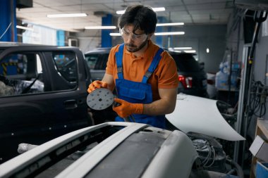 İşçi bir oto tamirhanesinde araç gövdesi paneli üzerinde yörünge zımparası kullanıyor, boya için yüzey hazırlıyor.