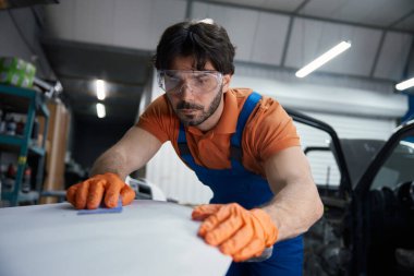 Teknisyen atölyede araba panelini zımparalıyor. Yetenekli işçiler güvenlik gözlüğü ve eldiven takıyor, yüzey hazırlıklarına odaklanmış..