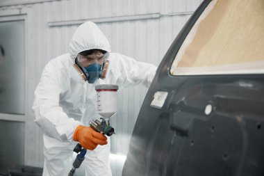 Koruyucu giysiler giyen bir teknisyen, otomotiv parçalarını dikkatlice sprey boyayla boyuyor.