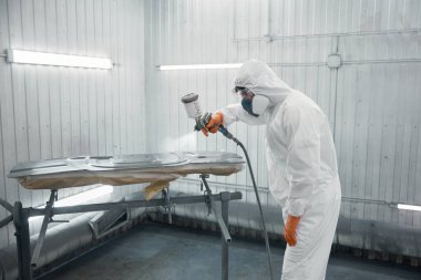 Tam korumalı ekipman teknisyeni havalandırmalı boya kabininin içine bir otomotiv paneli boyuyor. Arabanın bir parçası olan yetenekli bir işçi.