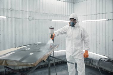 Koruyucu giysiler giyen endüstriyel ressam, temiz, havalandırmalı boya kabinindeki bir oto paneline sprey boya sürüyor..