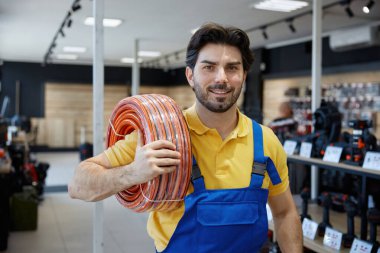 Mavi tulumla birlikte parlak sarı bir gömlek giyen bir adam elinde bir hortum tutuyor ve bir görev üzerinde çalışmaya hazır.