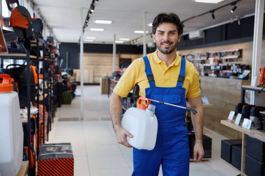 Mavi tulumlu bir adam perakende mağazasında duran bir sprey tutuyor ve müşterilere temizliği sürdürmeleri için yardım etmeye hazır.
