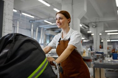 Pratik önlüklü bir kadın, iş görevinin bir parçası olarak büyük siyah çantalı bir arabayı iterek yere doğru ittiriyor.