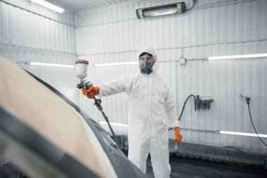 Otomotiv boya kabinine araba boyamak için sprey tabanca kullanan koruyucu kıyafetli bir işçi..