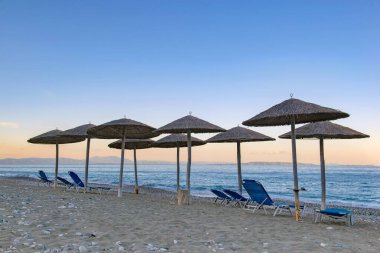 Mora Yunanistan 'da gün batımında bir plajda saman şemsiyesi ve güneş şezlongları