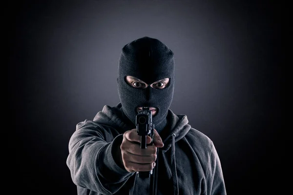Criminal wearing black balaclava and hoodie with a gun in the dark