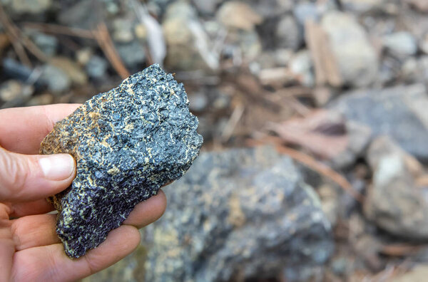 Fingers holding piece of chromite ore over natural rocks background, soft focus