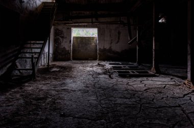 Eski fabrikanın arka planı. Yerde kırık toprak, kırık ahşap merdivenler ve paslı makine kalıntılarıyla dolu karanlık bir oda.