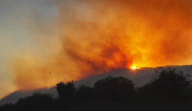 Gün batımında bir dağ yamacında şiddetli kırmızı gökyüzü, ağır duman ve su kovası ile helikopter silueti