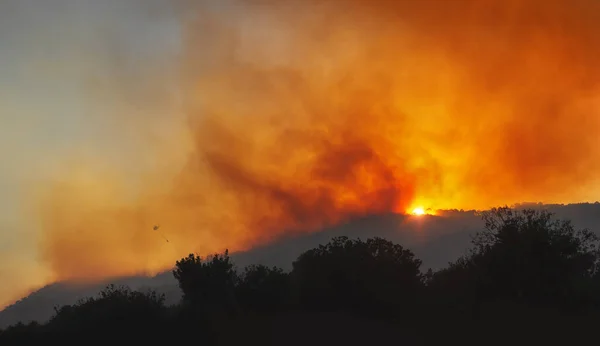 Wildfire on a mountain slope at sunset with dramatic red sky, heavy smoke and silhouette of helicopter with water bucket